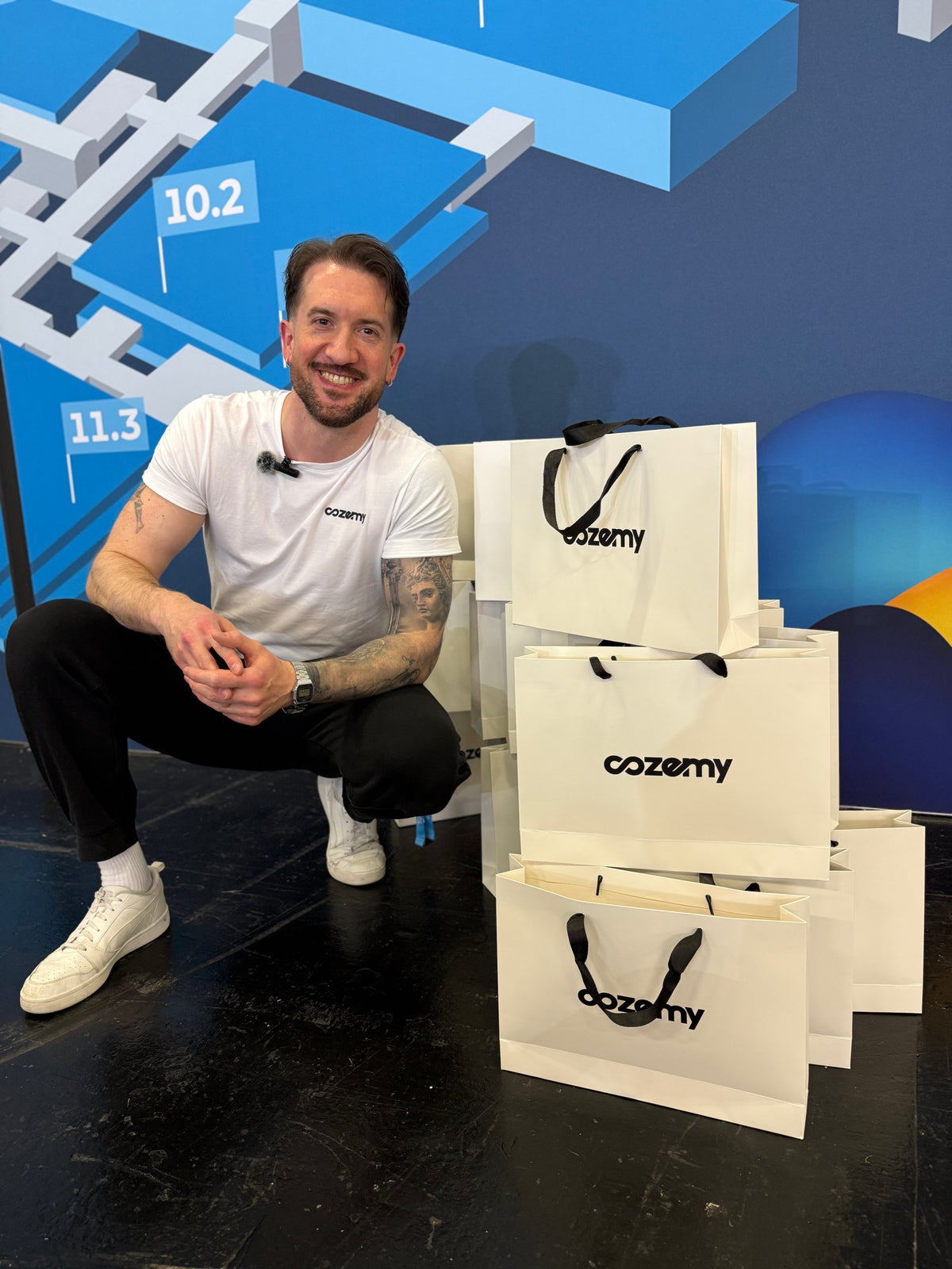 Lächelnder Mann im Cozemy T-Shirt hockt neben gestapelten Cozemy Papiertüten mit Logo